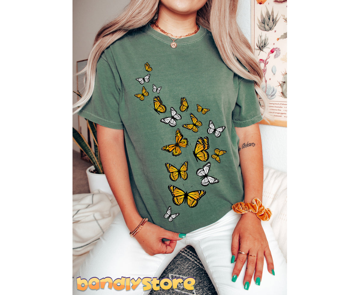 Monarch Butterfly Flock T-Shirt in moss on a seated person indoors near plants and a botanical poster.