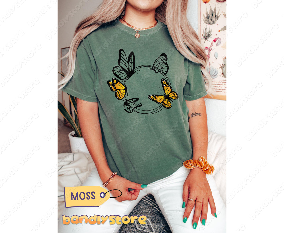 Monarch Butterfly Wreath T-Shirt in moss on a seated model facing forward amid indoor plants and wall art.