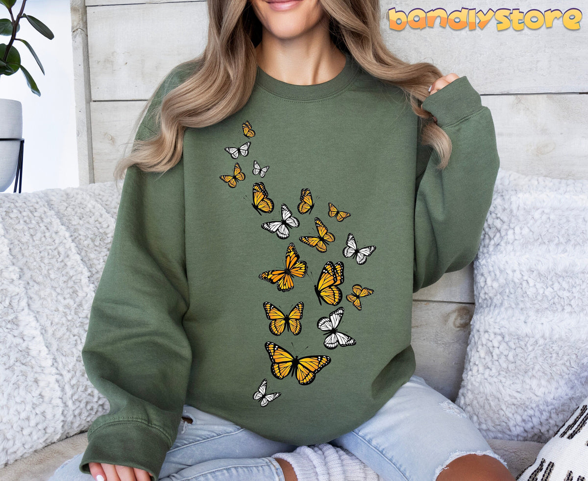 Monarch Butterfly Flock Sweatshirt in military green on a person seated on a white couch with pillows against a wooden wall.