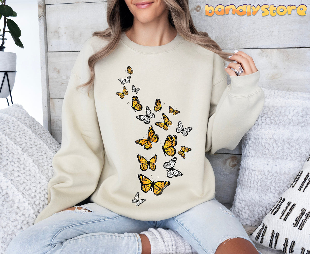 Monarch Butterfly Flock Sweatshirt in sand on a woman seated before a wooden wall with pillows and a plant.