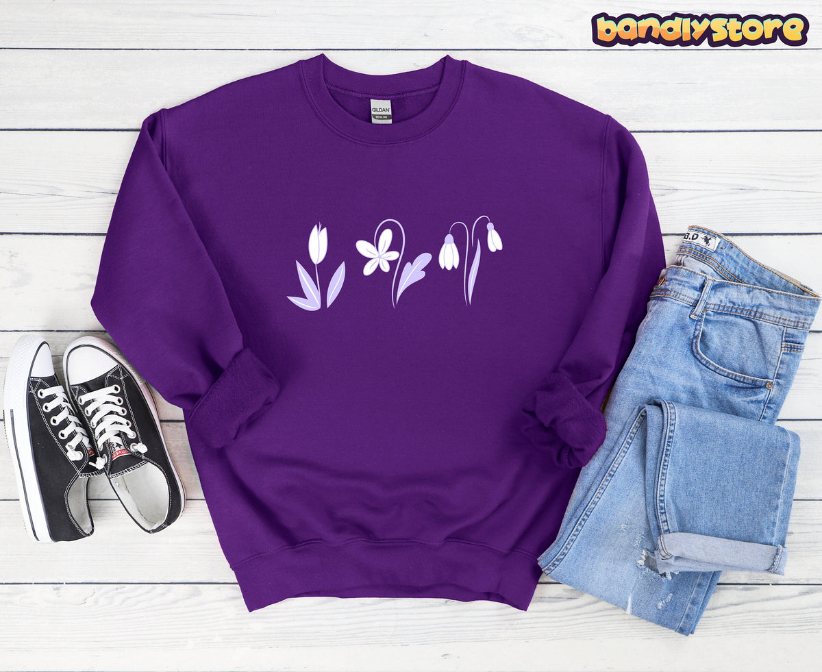 Monochrome Spring Flowers Sweatshirt in purple, flat-laid on white wood beside black sneakers and blue jeans.