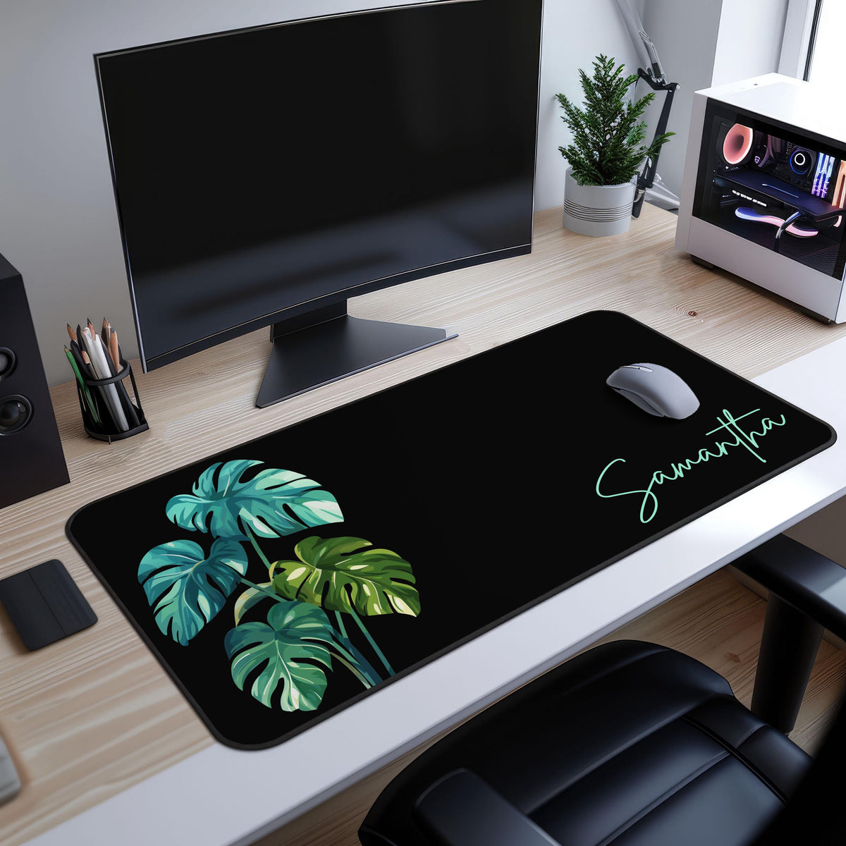 Personalized Monstera Desk Mat on a wooden desk under a monitor, with a mouse, potted plant and computer case nearby.