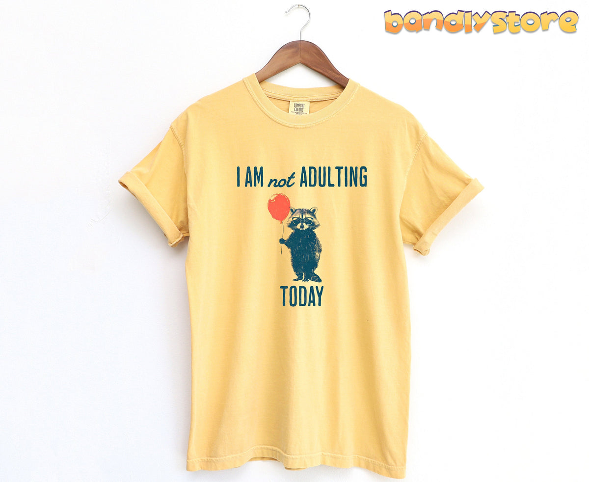 Not Adulting Raccoon T-Shirt in mustard hanging on a wooden hanger against a plain white wall.