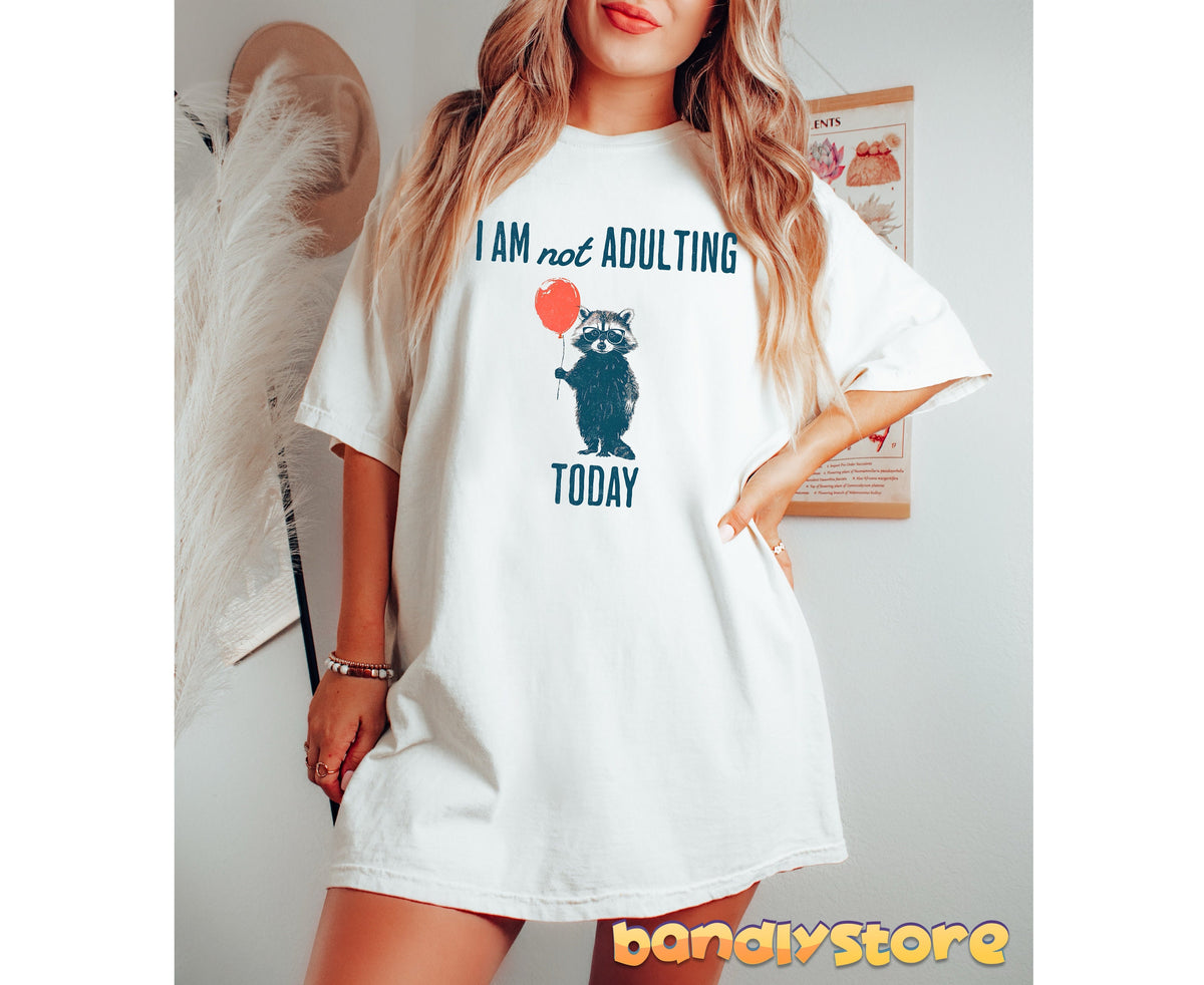 Not Adulting Raccoon T-Shirt in white worn by model standing indoors next to hat and botanical poster.