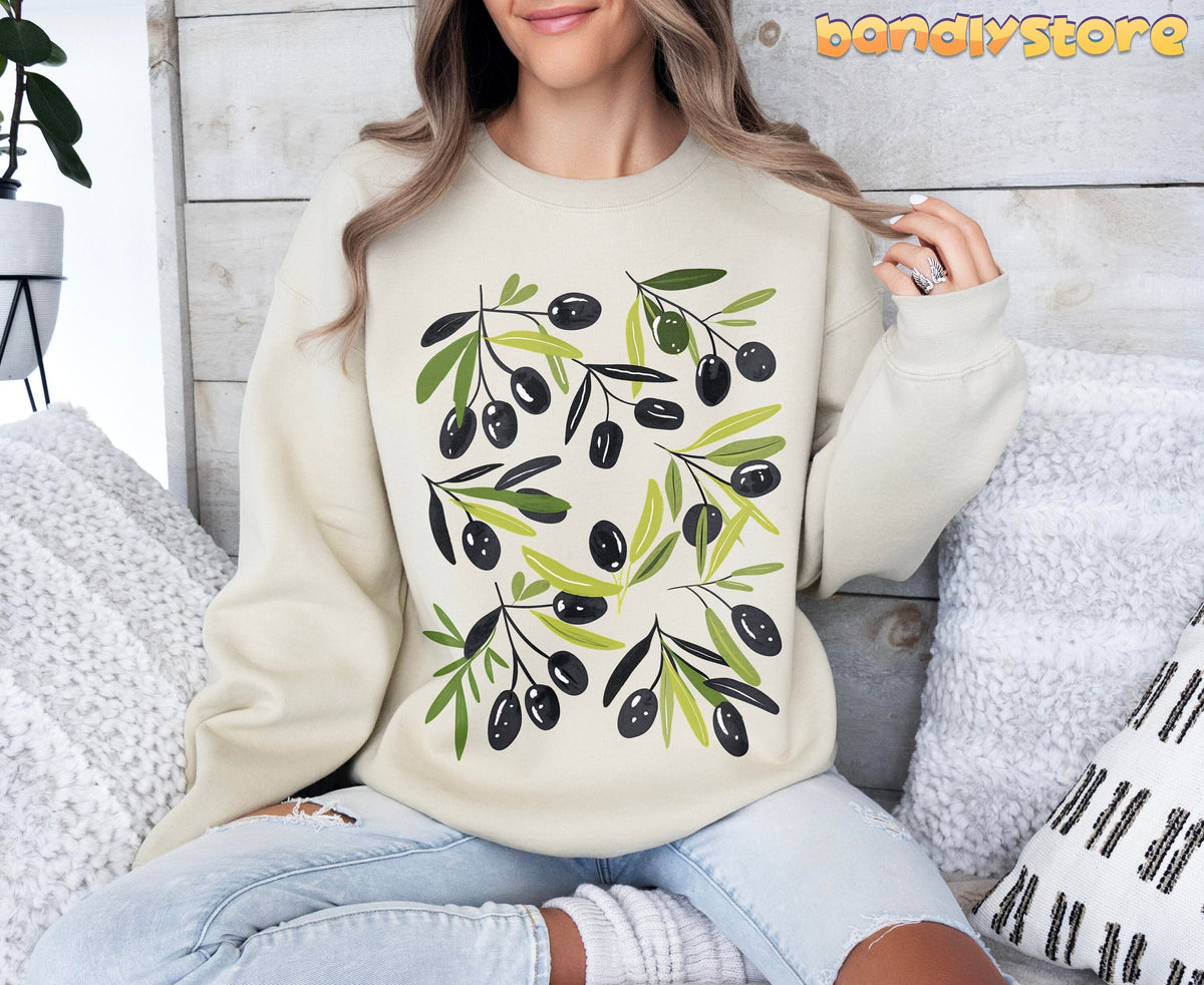 Olives Sweatshirt in sand on a seated woman against wood paneling with white pillows and a potted plant.