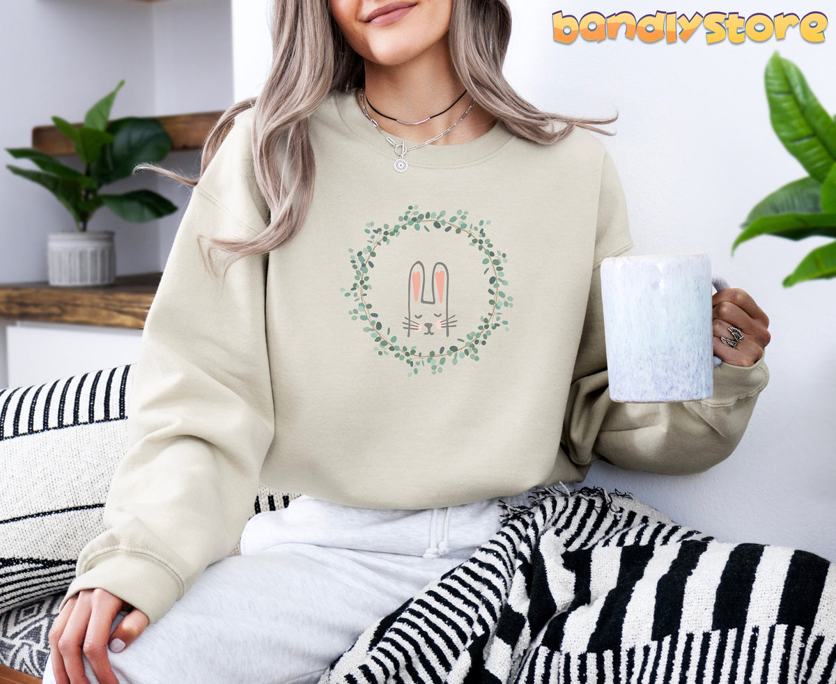 Spring Wreath Bunny Sweatshirt in sand, worn by a person seated indoors holding a speckled mug beside plants and striped textiles.