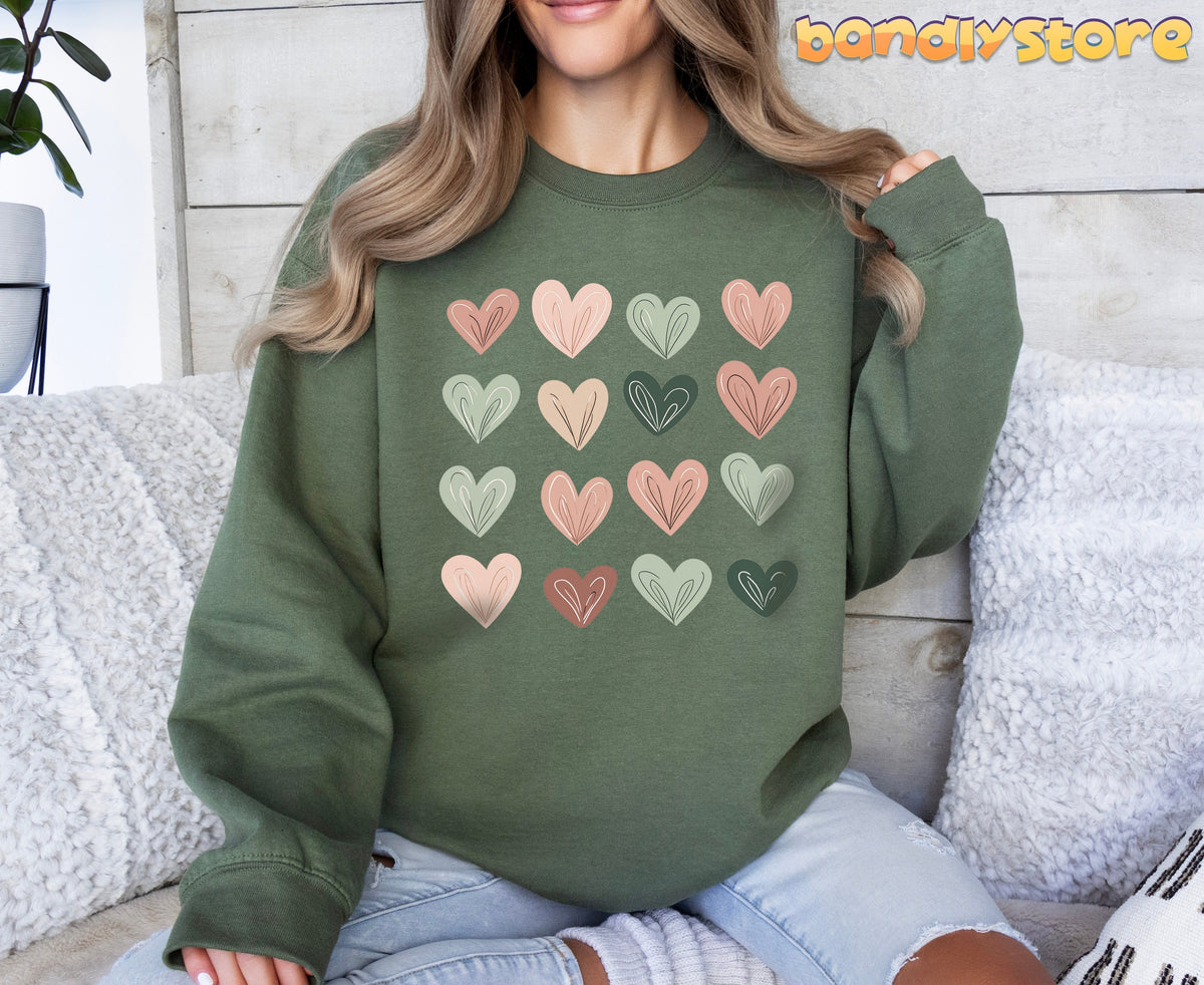 Cute Pastel Hearts Sweatshirt in military green on a person seated amid white pillows by a wooden wall and plant.