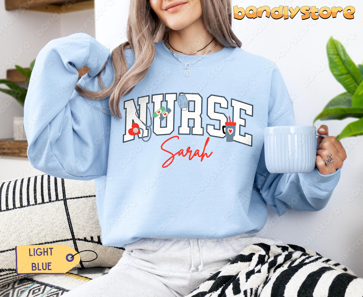 Personalized Nurse Sweatshirt in light blue worn by a woman holding a mug while seated on a couch beside plants.