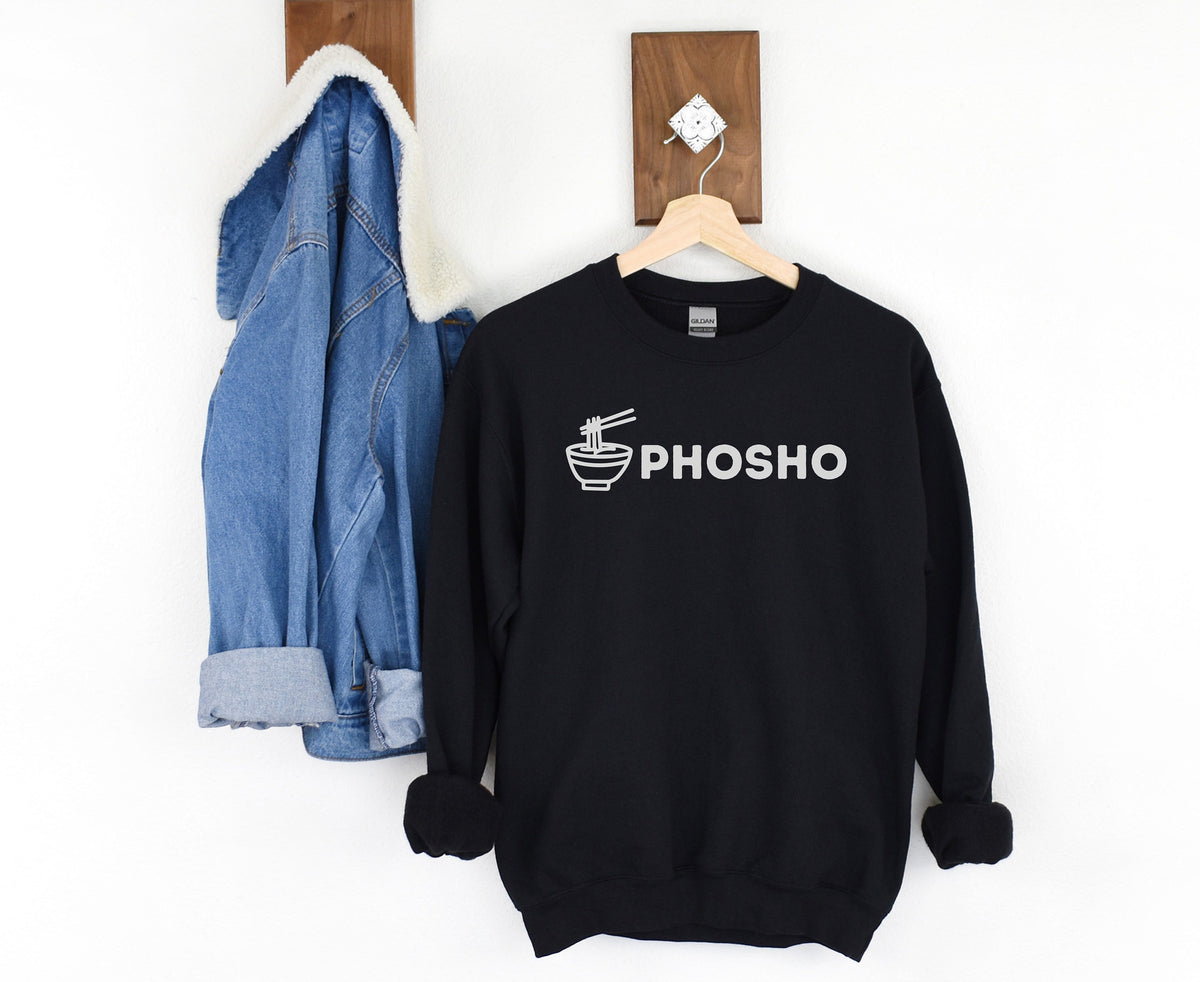 Pho Sho Sweatshirt in black hanging on a wooden hanger beside a denim jacket on a white wall.