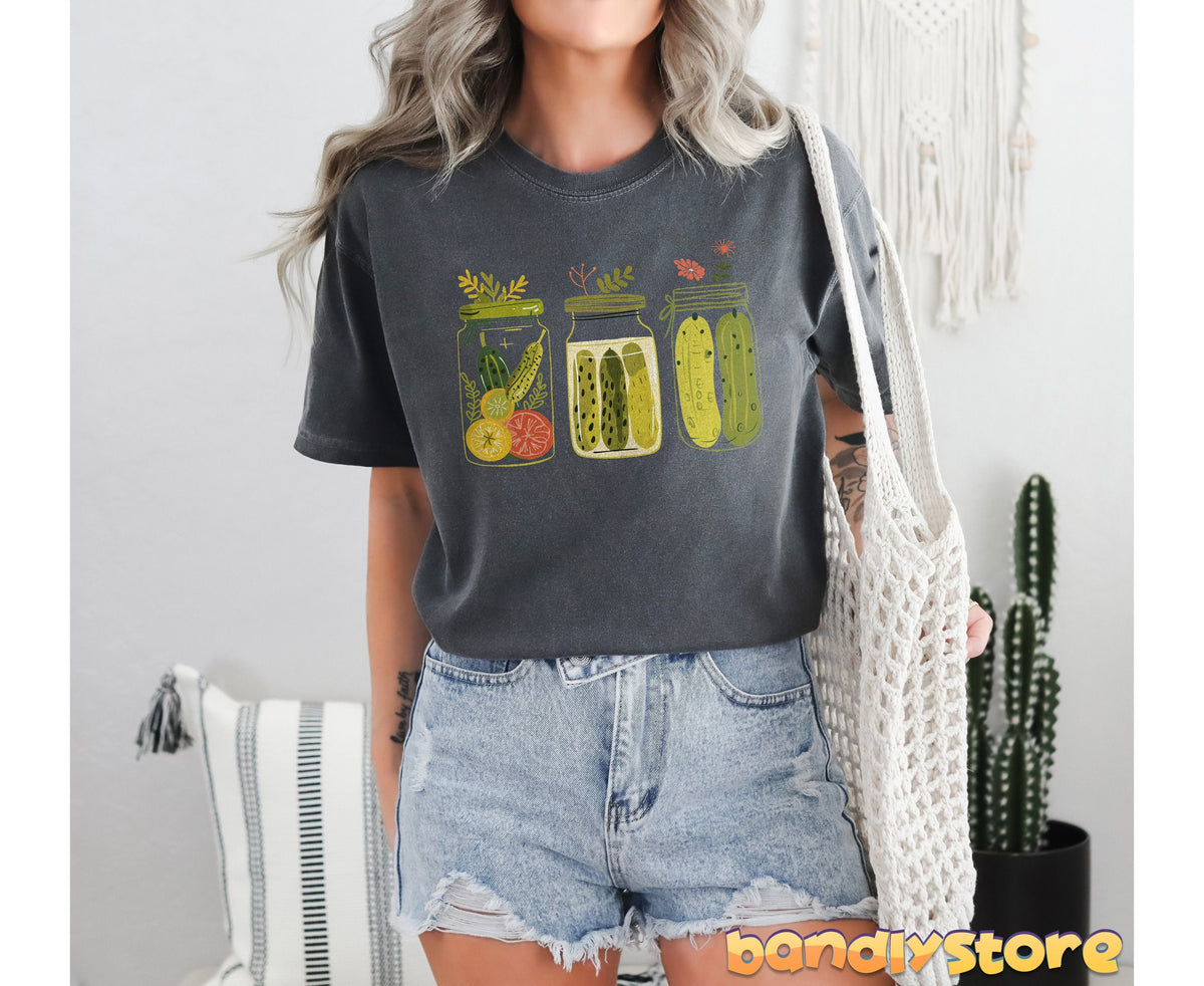 Pickle Jars T-Shirt in Pepper on a person standing indoors holding a net bag beside a cactus and pillow.