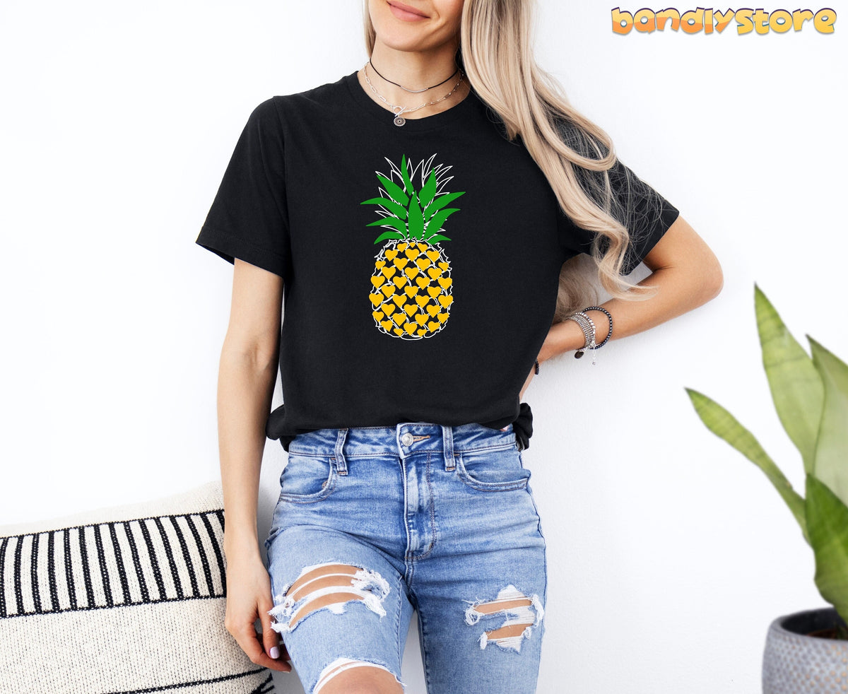 Pineapple T-Shirt on a black tee, woman standing by a white wall with plant and striped pillow nearby.