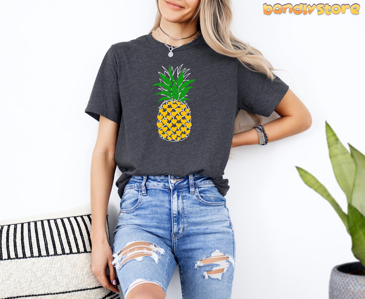 Pineapple T-Shirt in Dark Grey Heather on a model in front view against a white wall beside a striped pillow and plant.