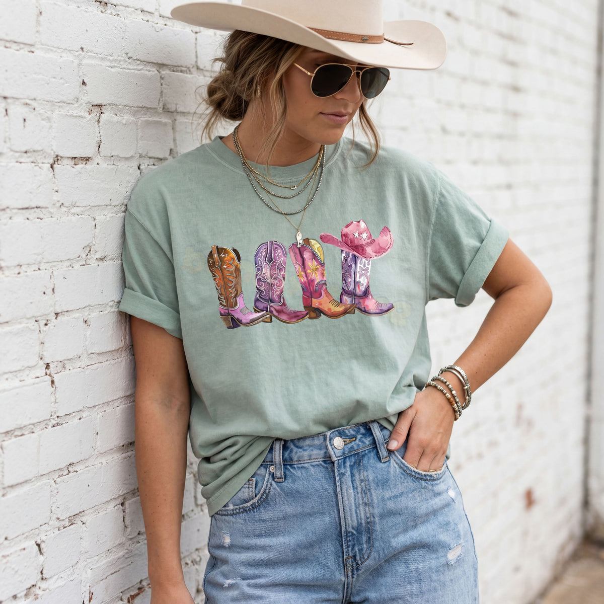 Pink Cowgirl Boots T-Shirt in Bay on a woman leaning against a white brick wall wearing a cowboy hat and jeans.