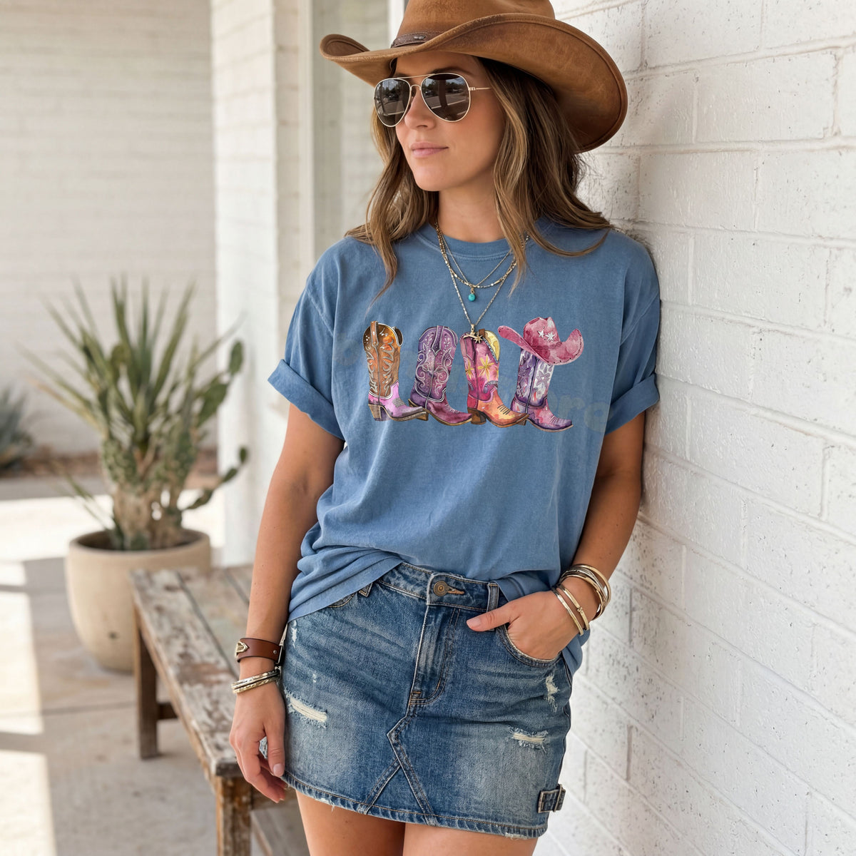 Pink Cowgirl Boots T-Shirt in Blue Jean worn by woman leaning on white brick wall beside potted plant and wooden bench.