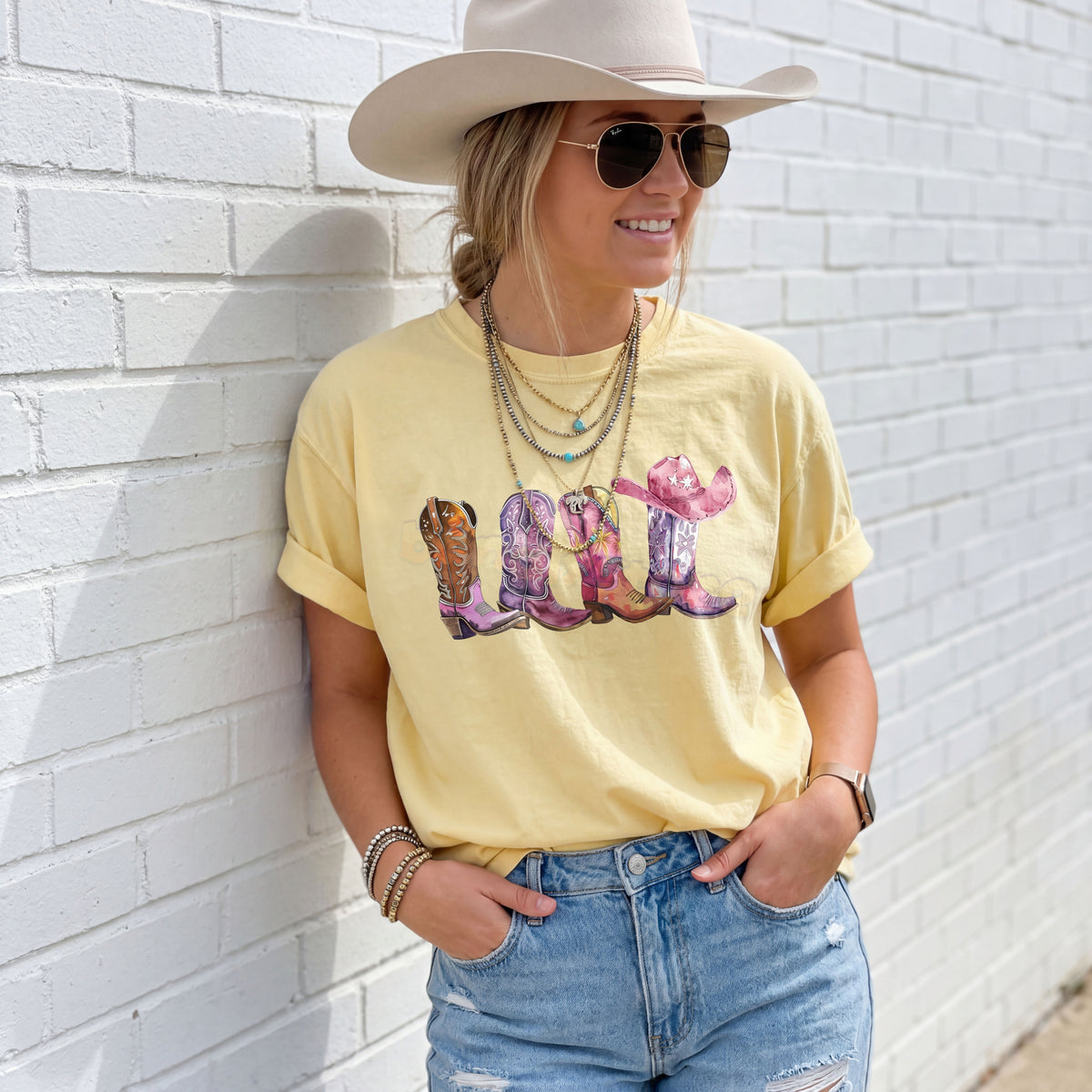Pink Cowgirl Boots T-Shirt in Butter, worn by a woman in a cowboy hat against a white brick wall.