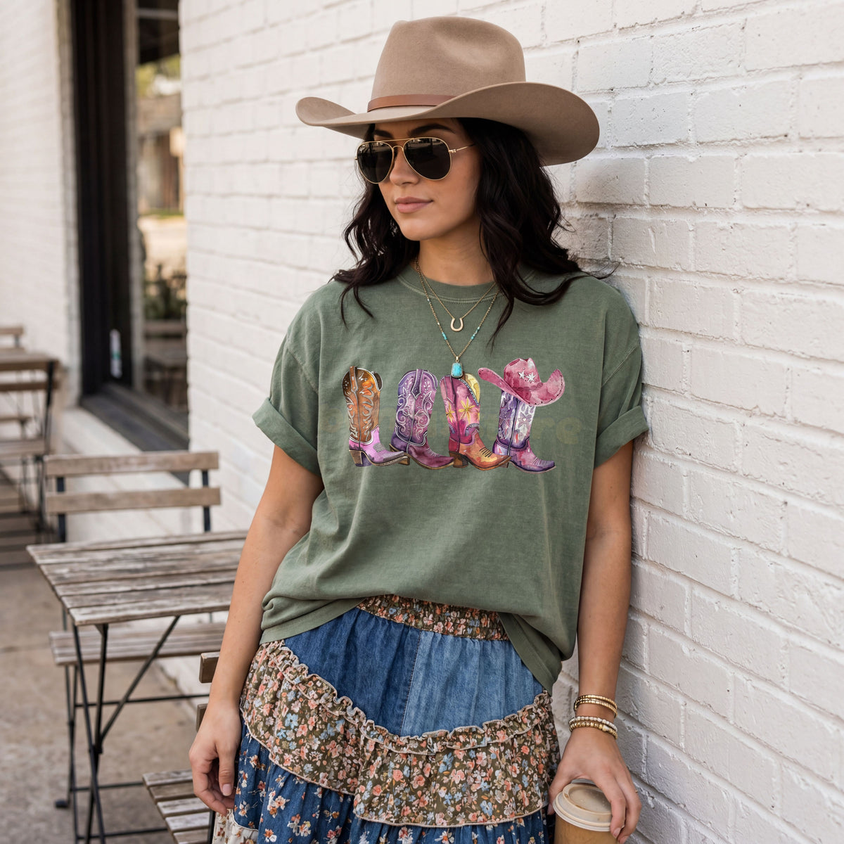 Pink Cowgirl Boots T-Shirt in moss on a woman leaning against a white brick wall outdoors beside tables, coffee cup in hand.
