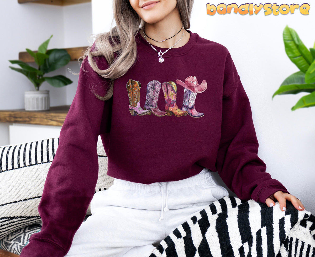 Pink Cowgirl Boots Sweatshirt in maroon, modeled seated indoors among plants and striped cushions.
