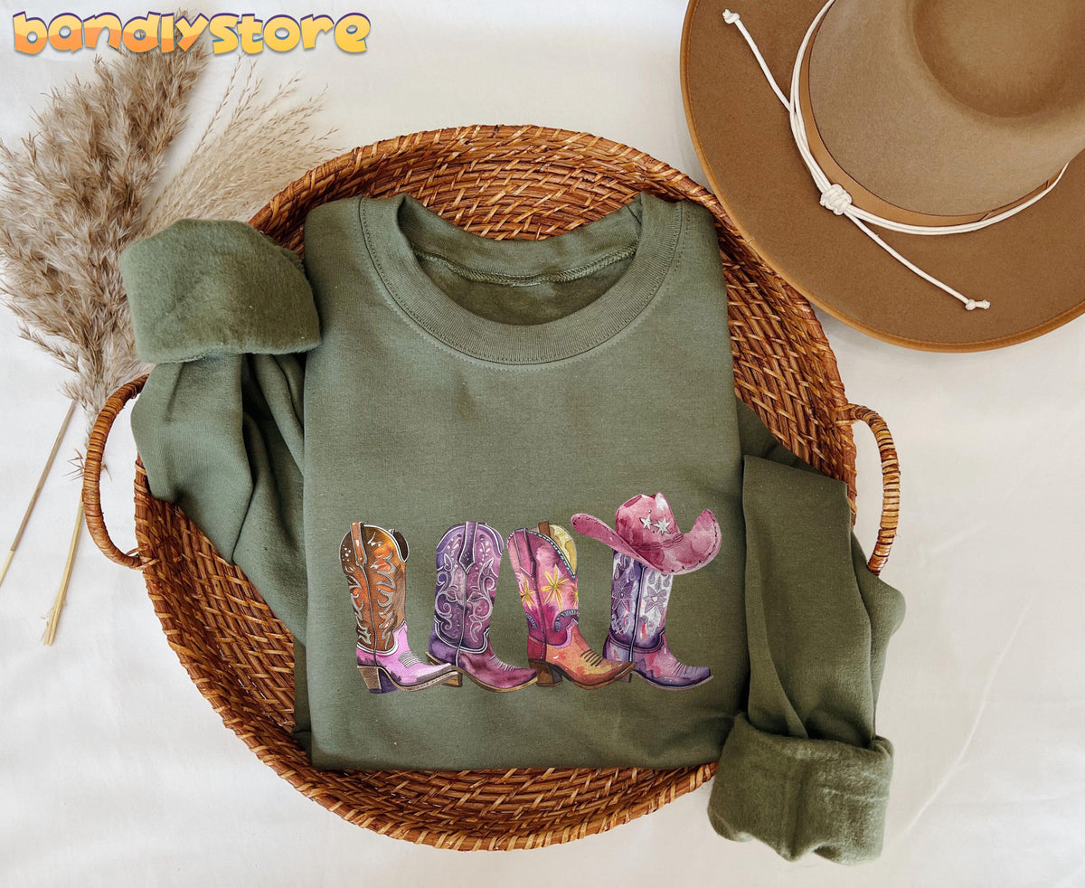 Pink Cowgirl Boots Sweatshirt in military green folded in a wicker basket beside a brown hat and pampas grass.