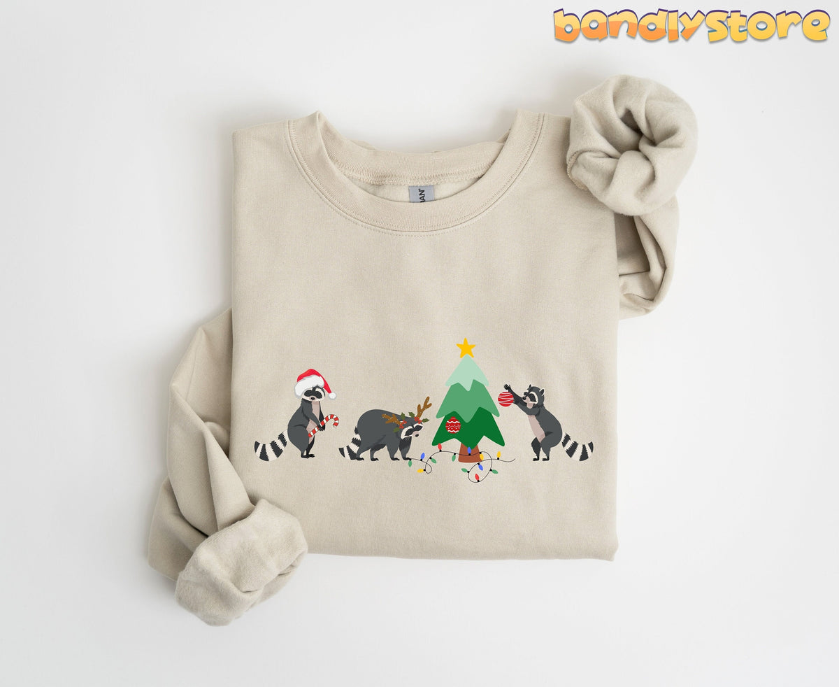 Raccoon Christmas Sweatshirt sand folded on white background with three raccoons decorating a Christmas tree.