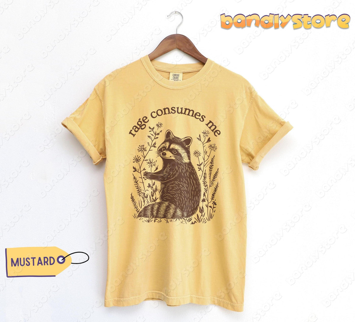 Rage Consumes Me T-Shirt in mustard hangs on a wooden hanger against a white wall with a raccoon graphic.