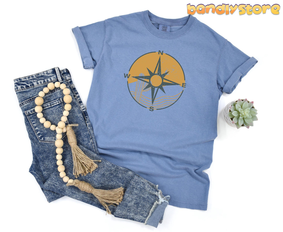 Retro Compass T-Shirt in Blue Jean laid flat beside jeans, wooden beads with tassels, and a small succulent.