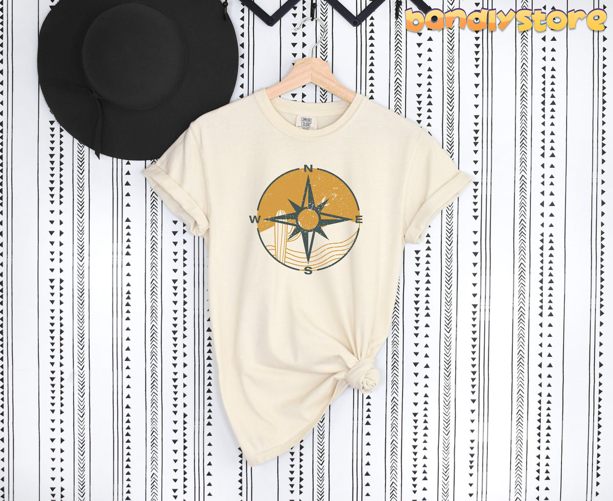Retro Compass T-Shirt in ivory, flat-laid on patterned wall beside black hat, knotted hem, wooden hanger.