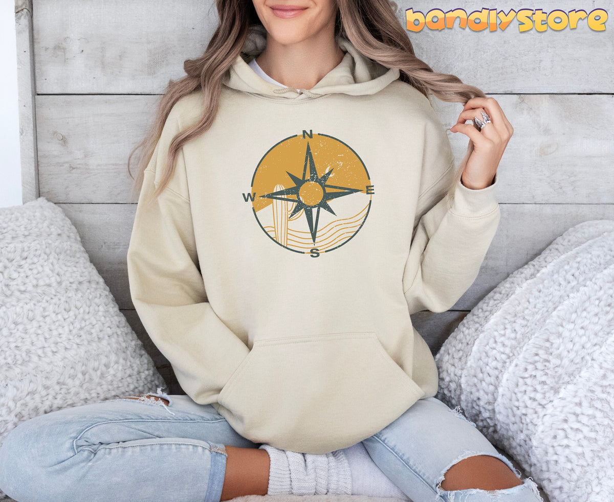 Retro Compass Hoodie in sand on a seated person against a wooden wall with white pillows.