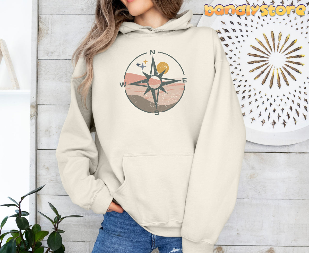 Retro Compass Hoodie in sand, front view on a person amid white wood walls, plants and a starburst decor.