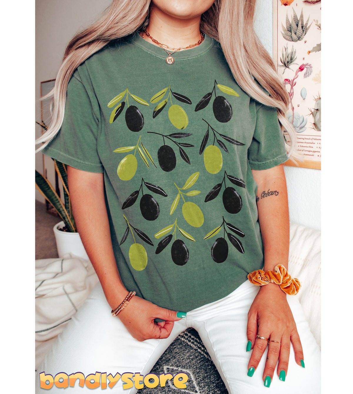 Retro Olives T-Shirt in moss on a seated person facing forward amid indoor plants and a botanical poster.