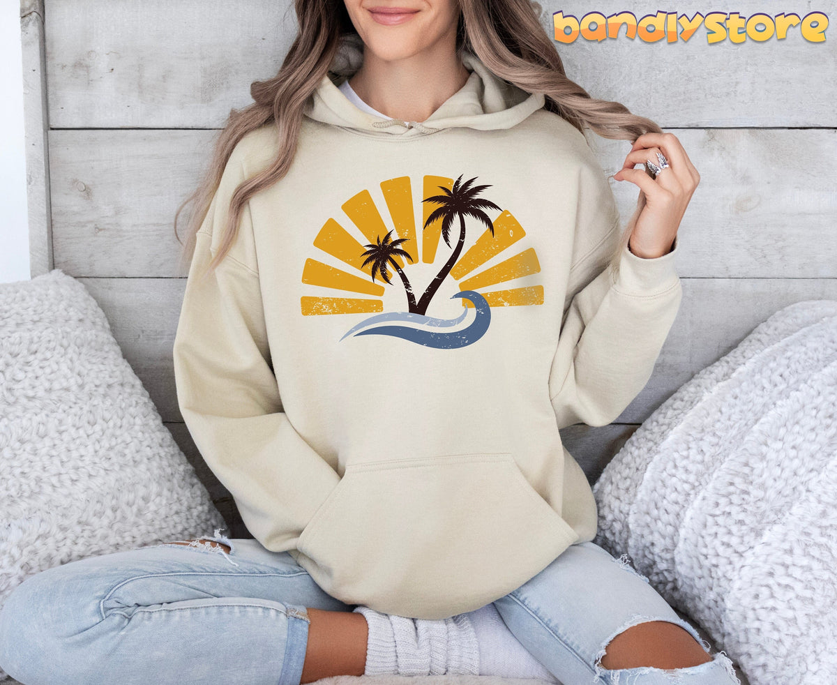 Retro Sunset Hoodie in sand worn by a person sitting cross-legged amid white knit pillows against a wood wall.