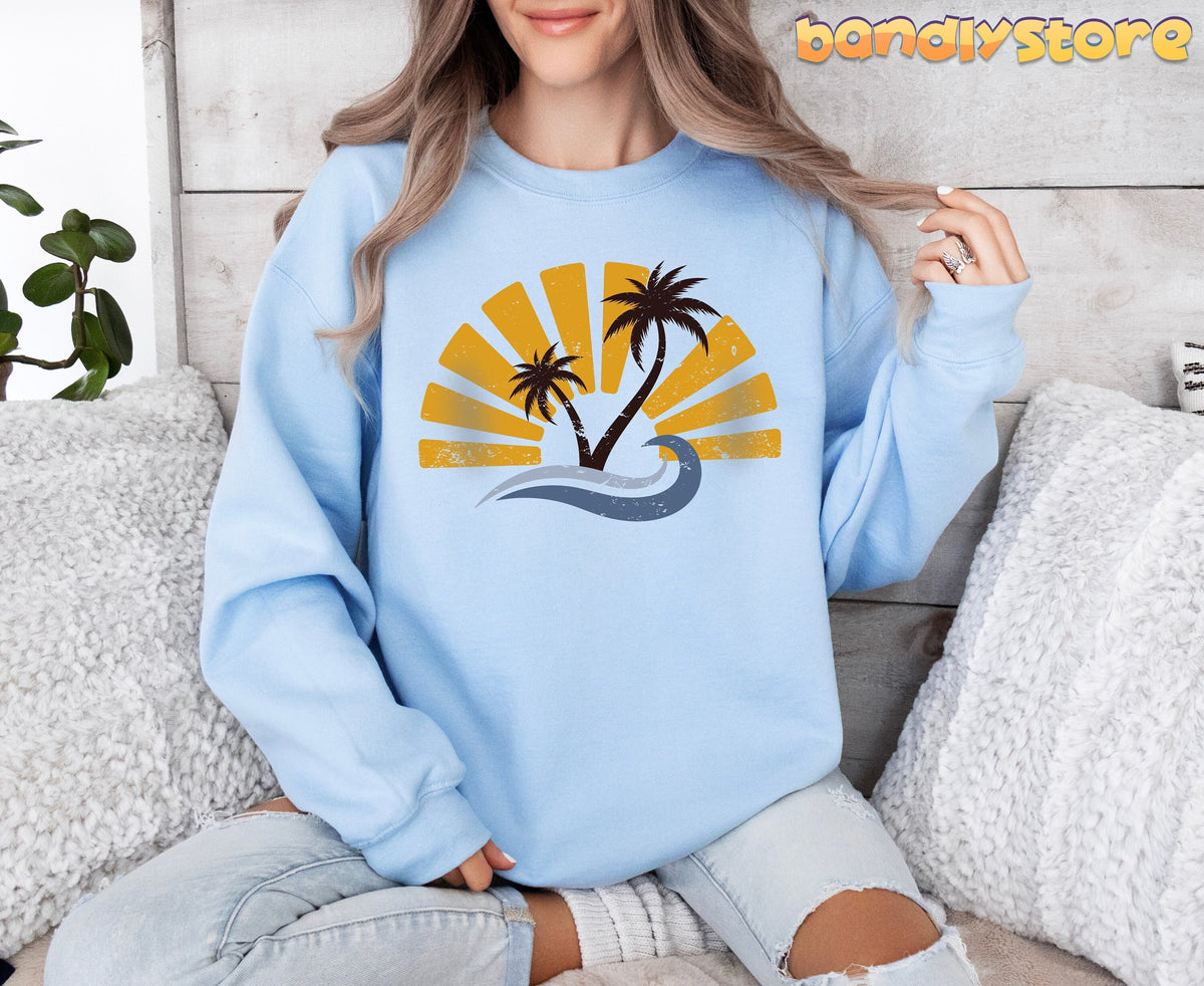 Retro Sunset Sweatshirt in light blue, worn by a seated person amid pillows and a plant against a wooden wall.