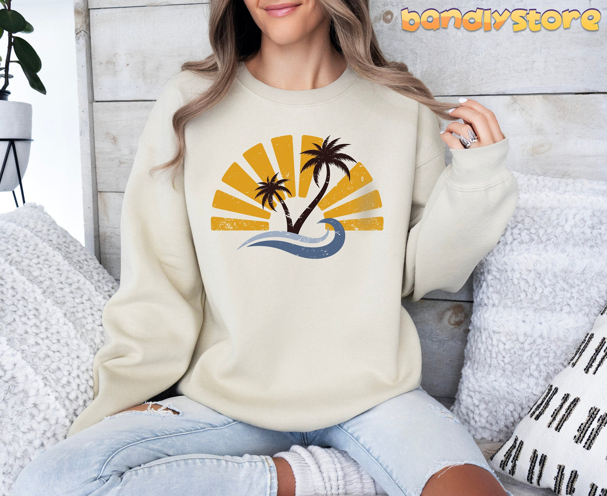 Retro Sunset Sweatshirt in sand, worn by a seated woman among pillows against a wooden wall.
