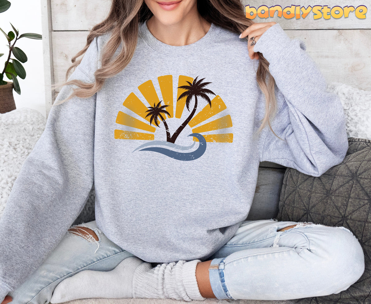 Retro Sunset Sweatshirt in Sport Grey, worn by a seated person amid pillows and a plant by a light wood wall.