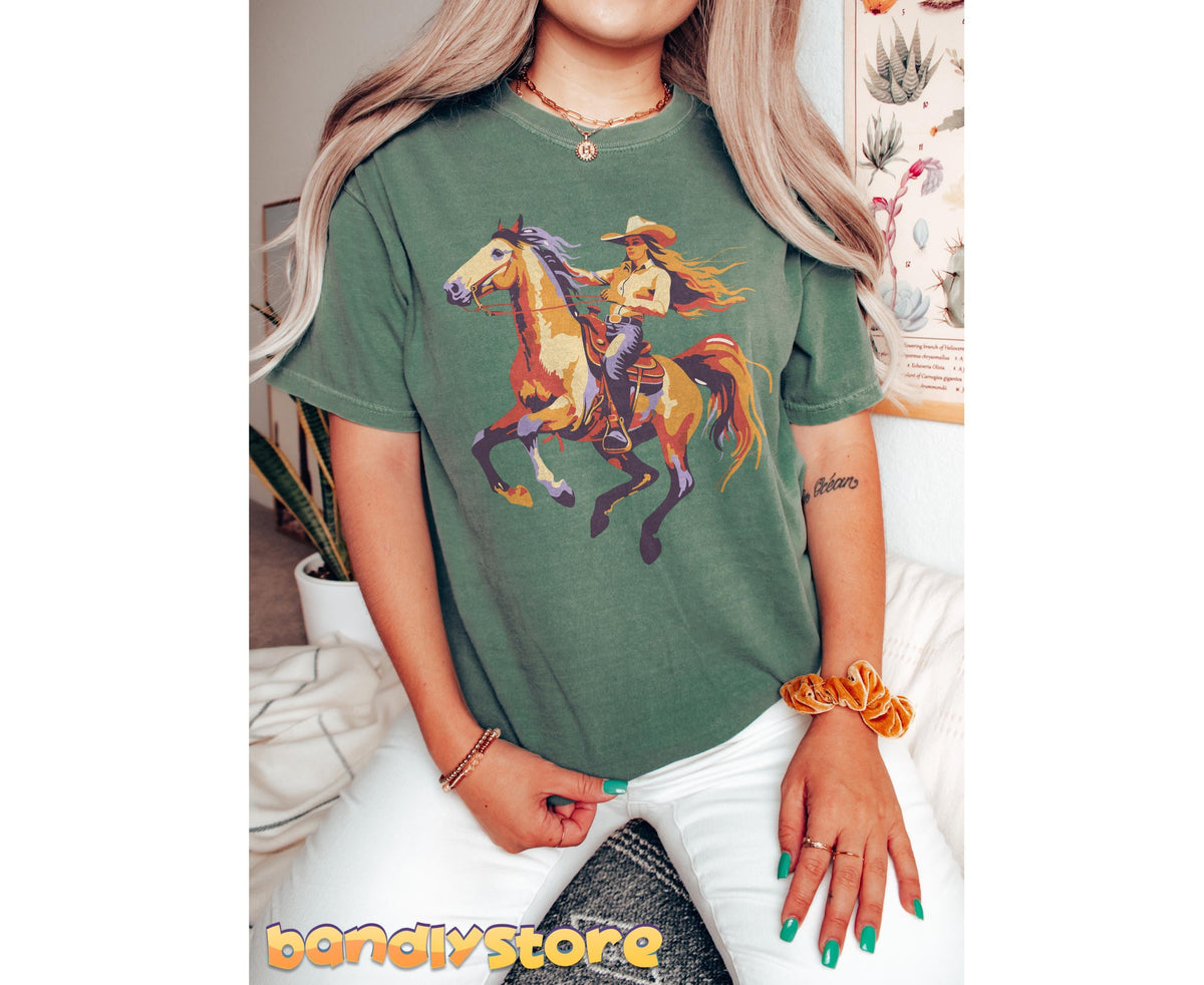 Retro Yeehaw Cowgirl T-Shirt in moss, worn front-facing with cowgirl-on-horse graphic amid plants and poster.