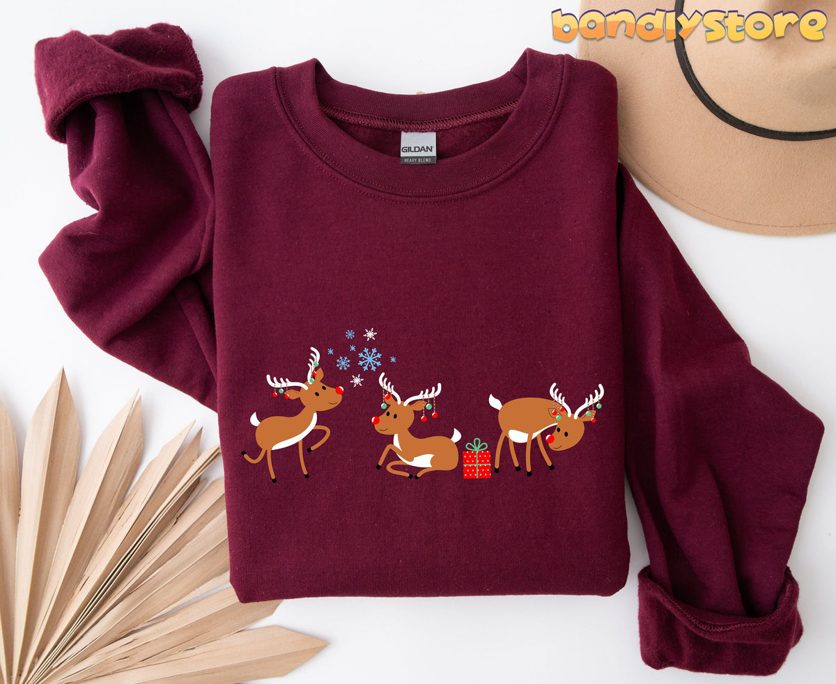 Rudolph The Red Nosed Reindeer Sweatshirt maroon folded flat with cartoon reindeer, snowflakes, gift, hat and dried leaves nearby.