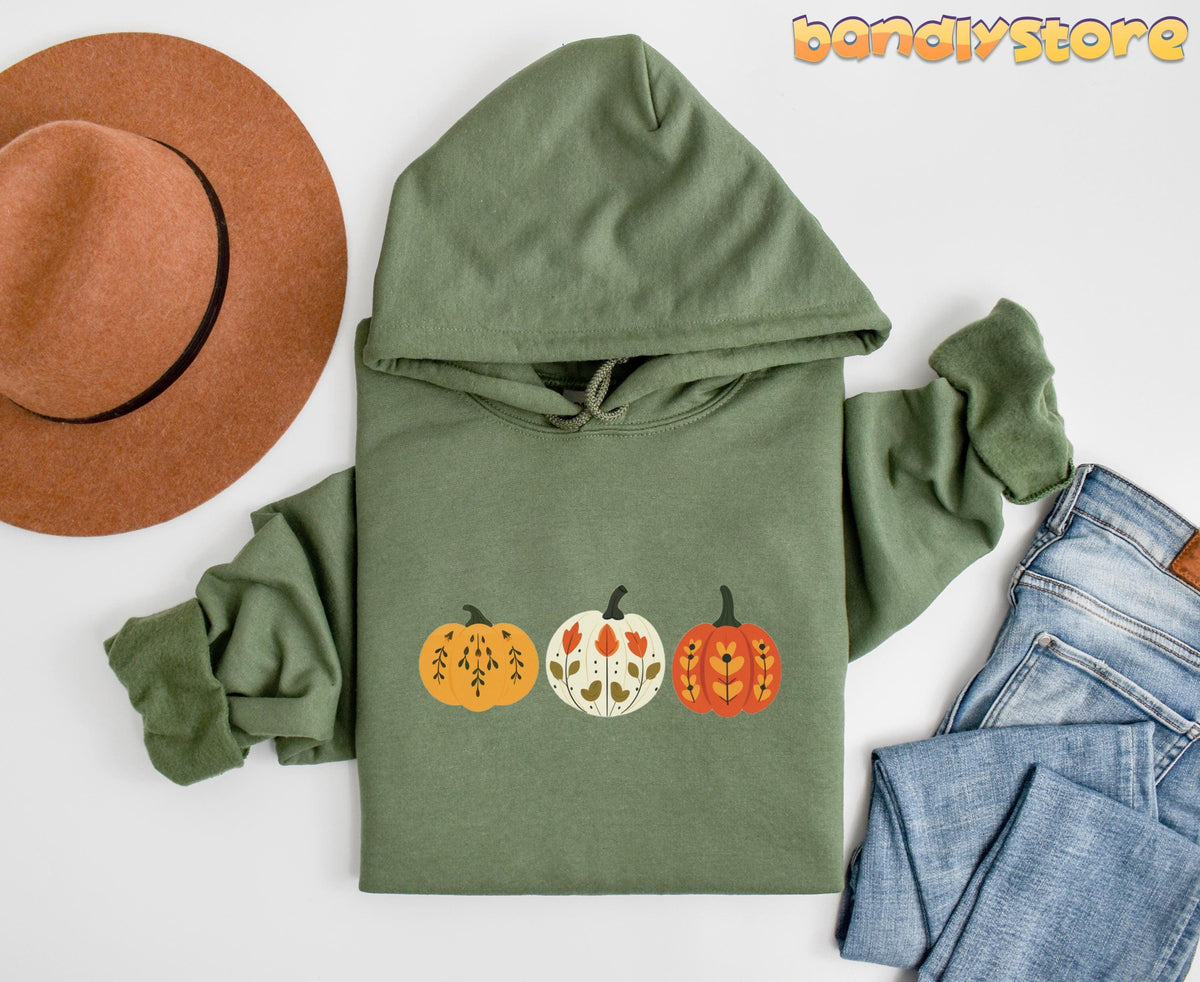 Scandinavian Art Pumpkin Hoodie in military green, flat-lay with brown hat left and blue jeans right.