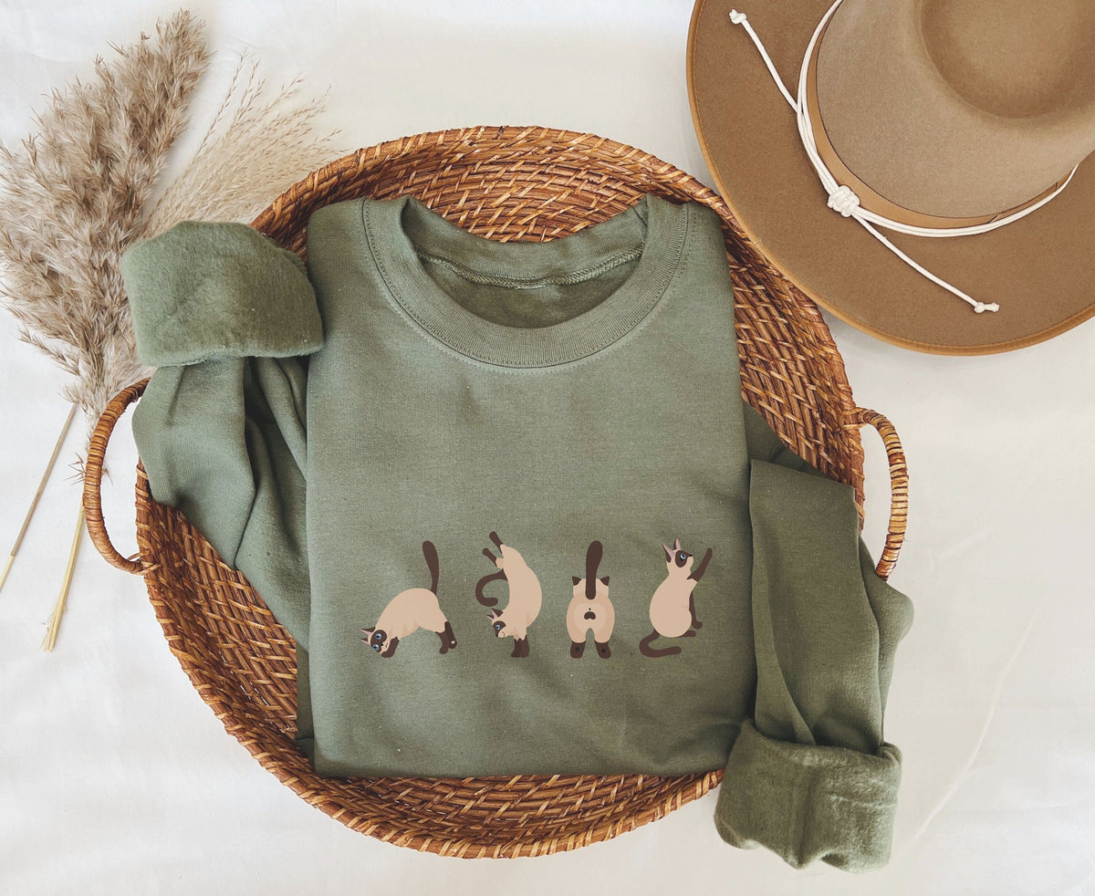 Siamese Cat Butts Sweatshirt in military green folded inside a wicker basket beside a hat and dried grass, top-down view.