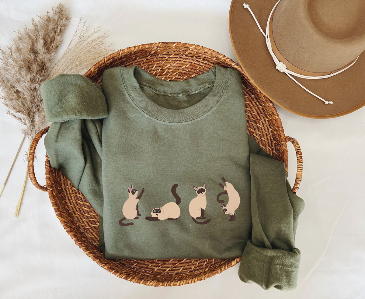 Siamese Cat Sweatshirt in military green, folded in a woven basket beside a hat and dried grass.