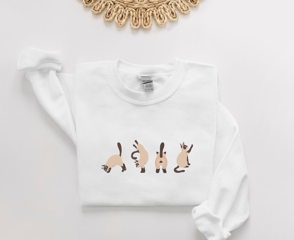 Siamese Cat Butts Sweatshirt in white laid flat on a white surface below a woven rattan piece.