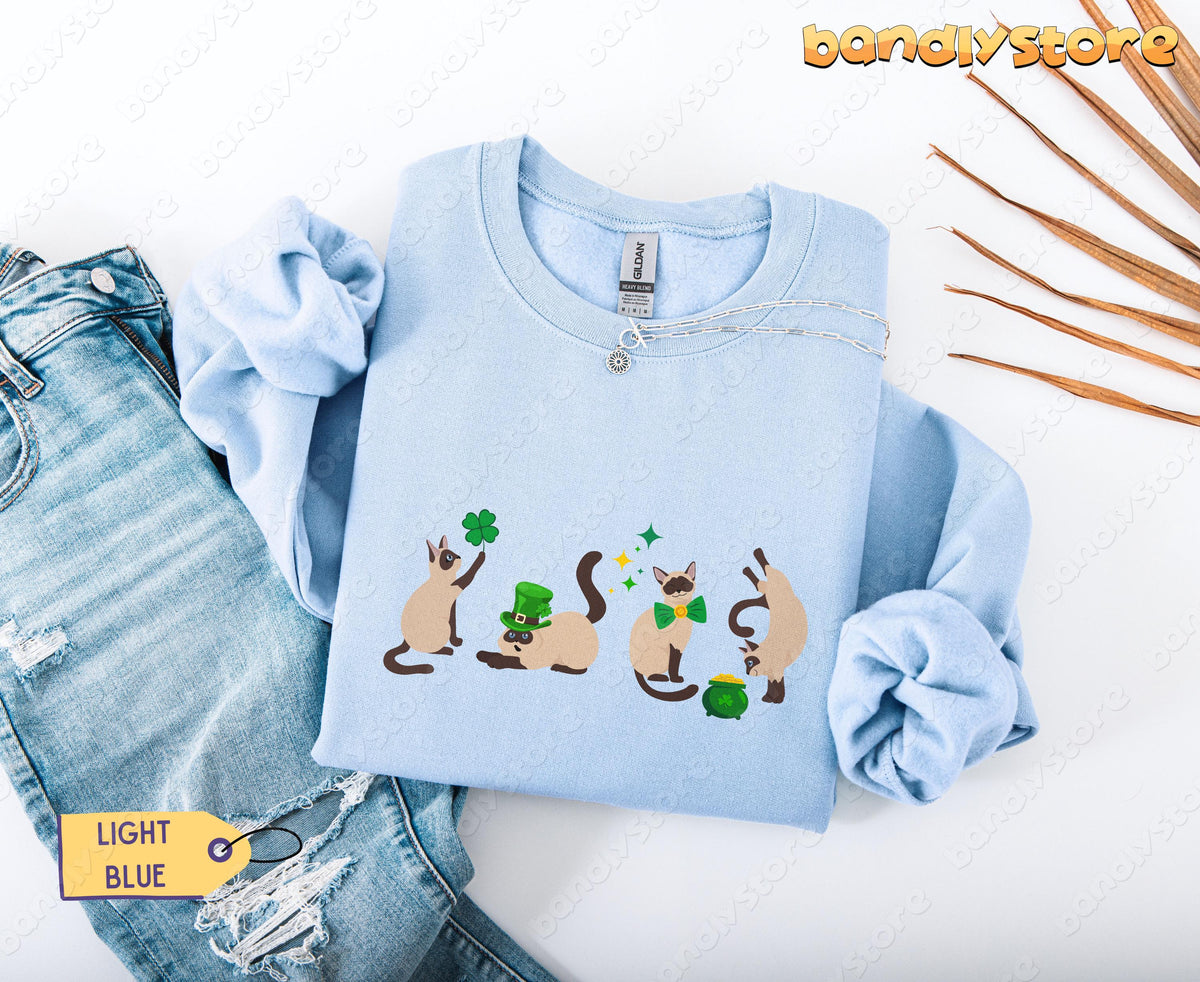Siamese Cat St Patrick's Day Sweatshirt in light blue, folded flat beside ripped jeans, necklace and dried palm leaves.