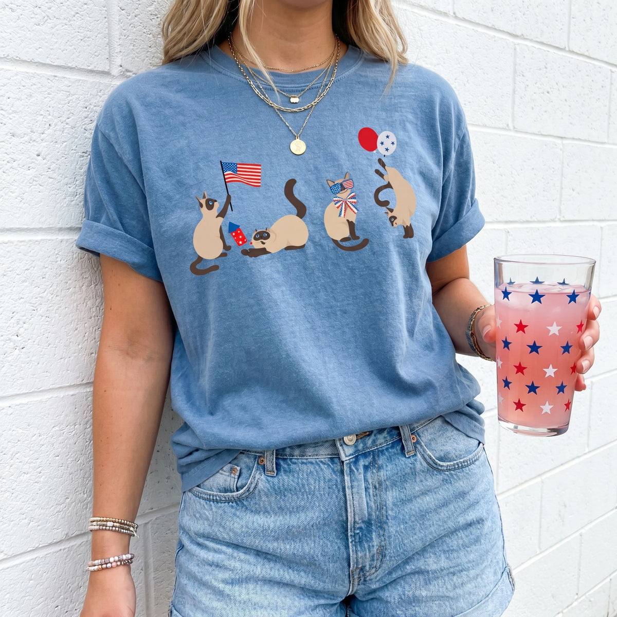 Siamese Cats 4th of July T-Shirt in Blue Jean, front view on a person by a white brick wall holding a star glass.