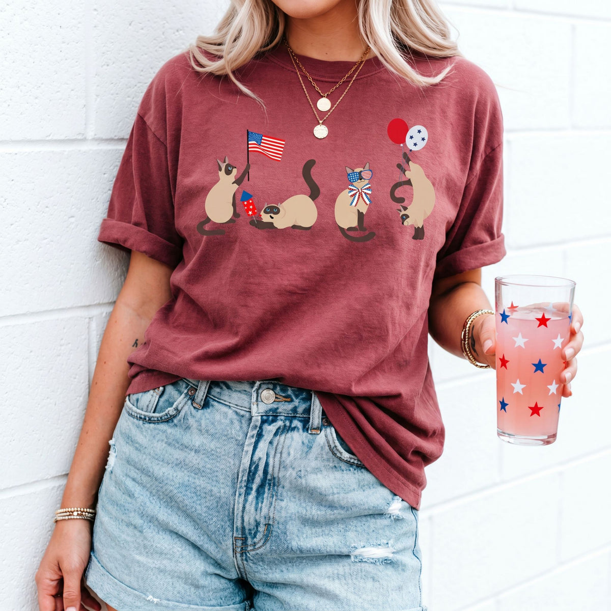 Siamese Cats 4th of July T-Shirt in brick on person against white brick wall holding starred glass.