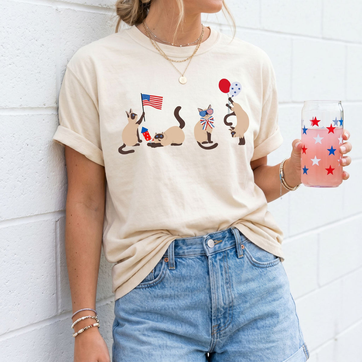 Siamese Cats 4th of July T-Shirt in ivory, worn front-view against a white brick wall with a starry drink.