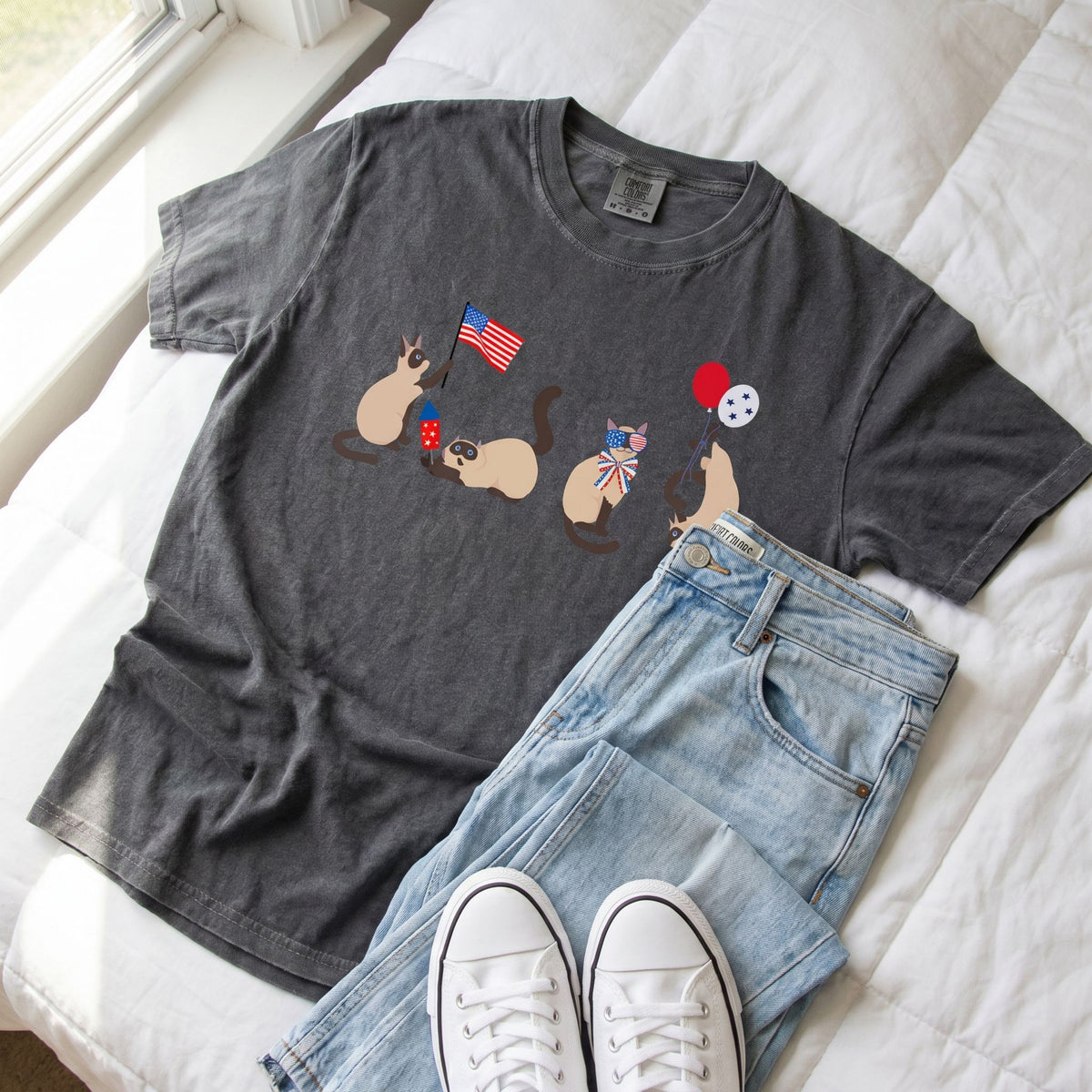 Siamese Cats 4th of July T-Shirt in pepper laid flat on white bedding beside jeans and white sneakers.