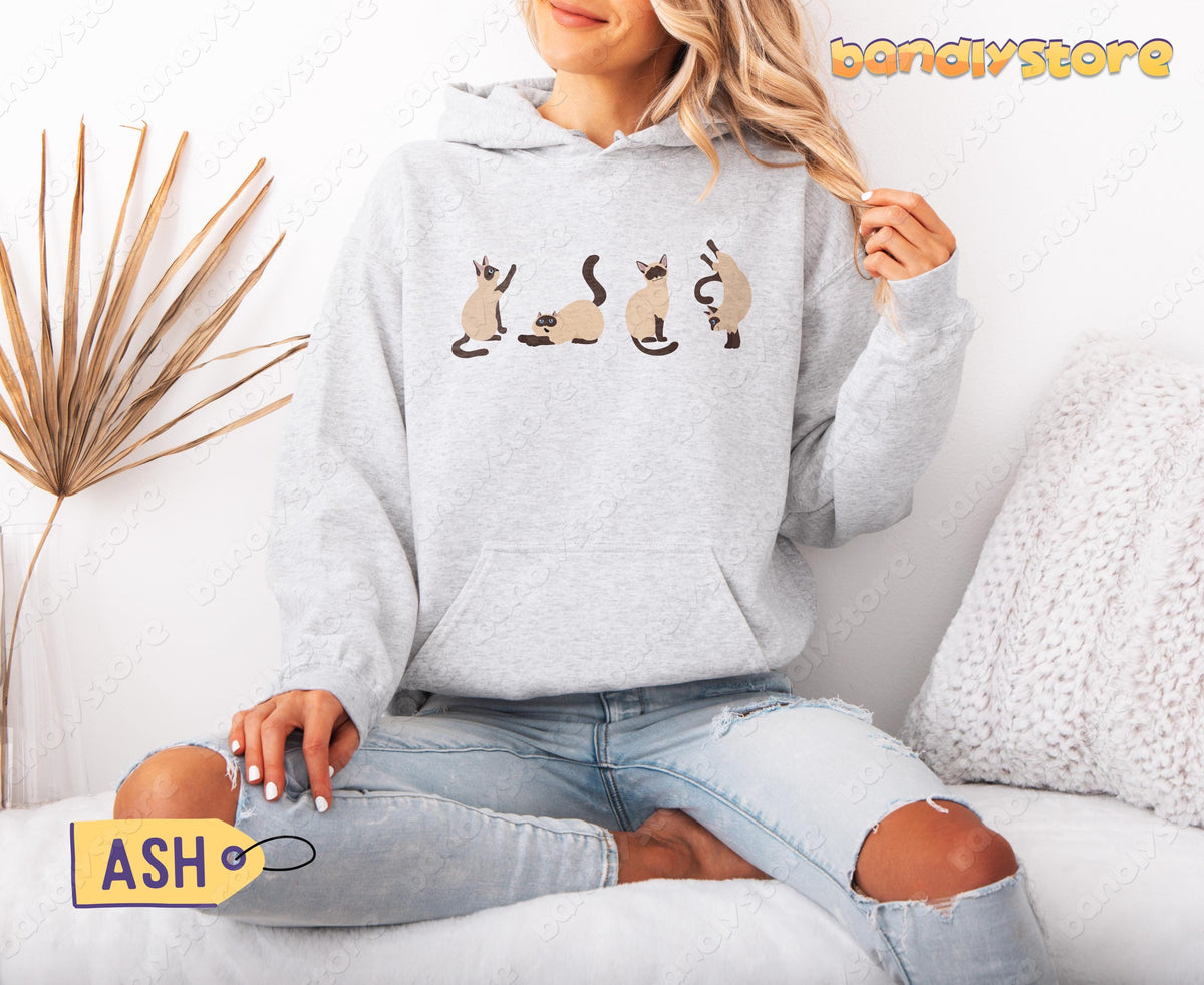 Siamese Cats Hoodie in ash, front view on a model sitting cross-legged beside a dried palm frond and white pillow.