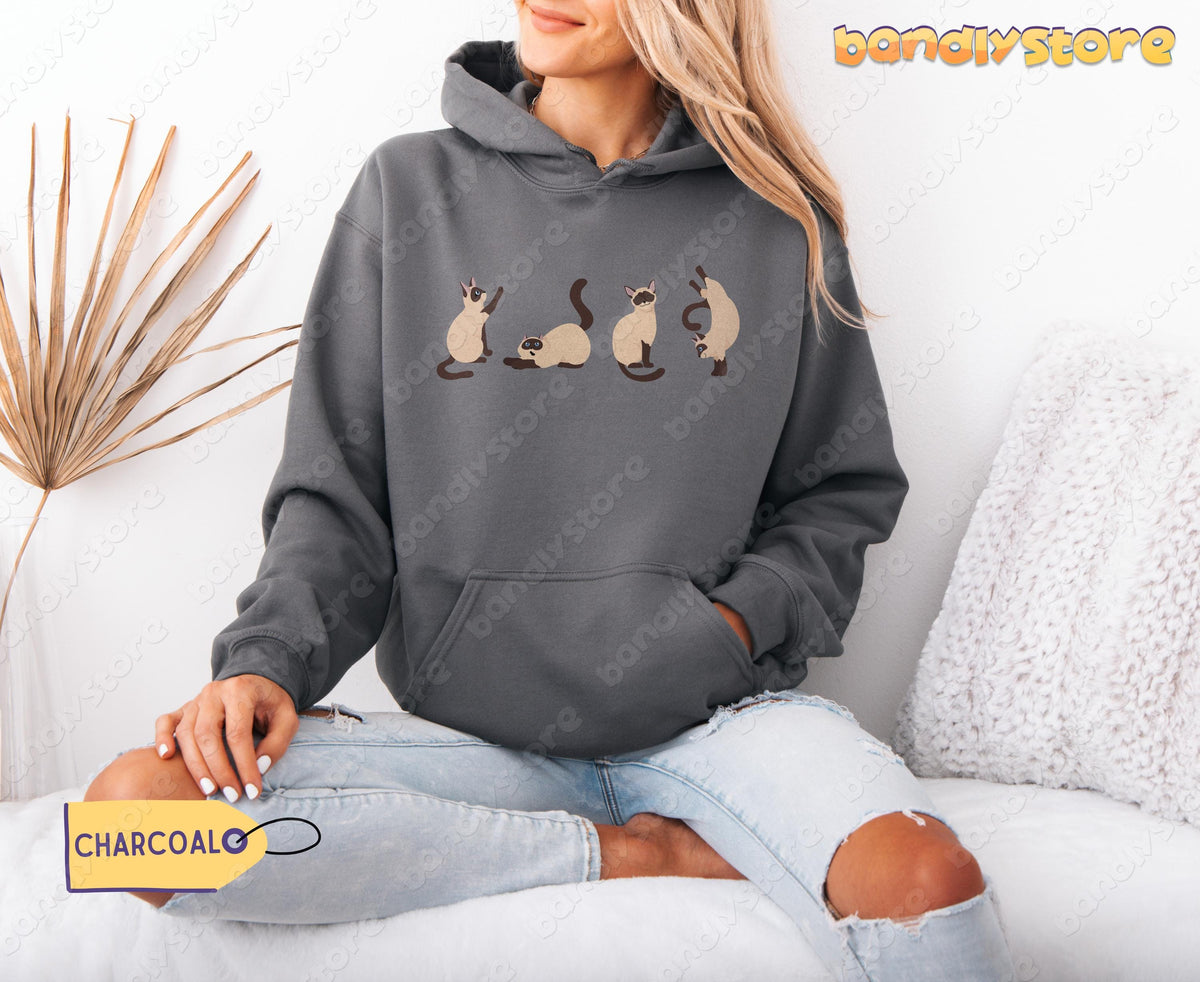 Siamese Cats Hoodie in charcoal on a person sitting cross-legged beside a dried plant and white pillow.