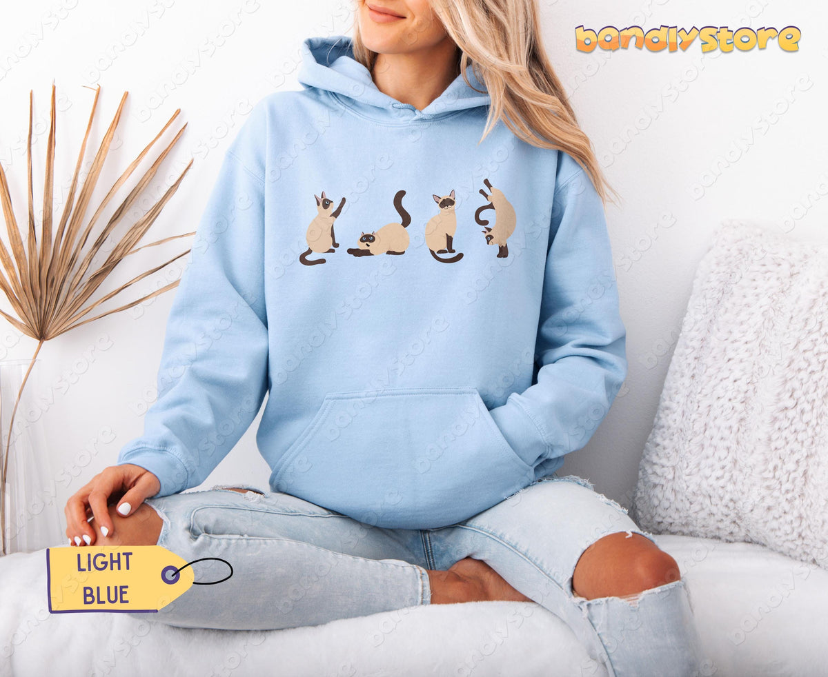 Siamese Cats Hoodie in light blue on a seated person beside a dried plant and cushion.