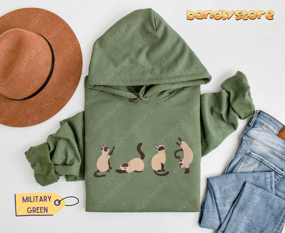 Siamese Cats Hoodie in military green flat-laid from above beside a hat and jeans on white.