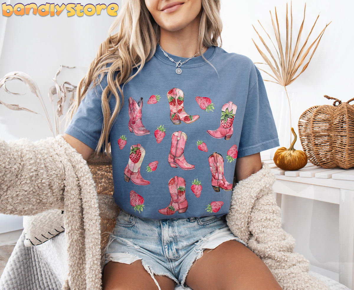 Strawberry Cowgirl Boots T-Shirt in Blue Jean worn by a seated model beside dried plants, a pumpkin and woven basket.