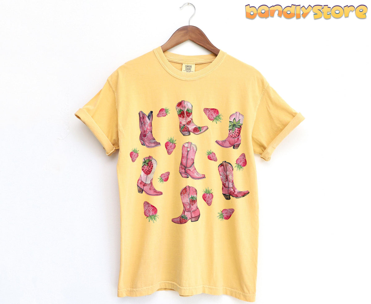 Strawberry Cowgirl Boots T-Shirt on a wooden hanger against a white wall, mustard tee with pink boots and strawberries.